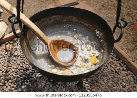 Left over food is at the bottom of a metal pan, hanging over a fire, with a wooden spoon in the pan.