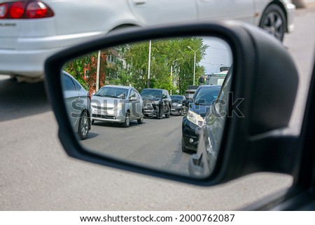 The left side black mirror of a passenger car, which reflects the cars behind standing in two rows on the road during the day.