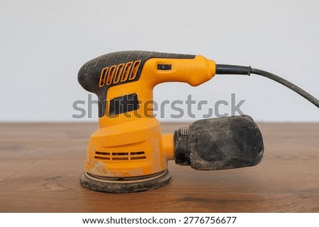 Left side view of a yellow orbital sander with a dust collection bag on a wooden background.
