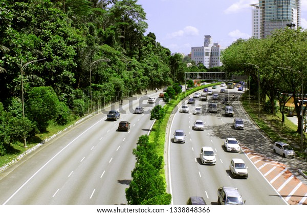 Left-hand traffic. Сars and motorcycles drive\
along a wide highway on a sunny day in a megapolis (big city) among\
tropical trees. Singapore view from\
above