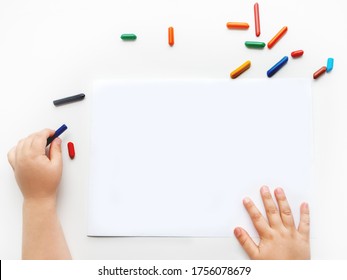 Left-handed toddler prepare to draw something on clear white sheet of paper. Kid uses wax crayons. Top view on child's hands and pencils on white background.