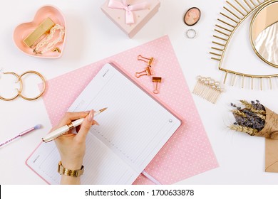 Left-hander girl keeps a diary and planning a day on stylish workplace. Female hand write in pink notebook. Women blog concept with pink notebook and golden watch. Flat lay style. 