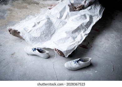 Leg with bare foot dead body lower part in sitting position was wrapped by white sack with dropped of blood and blurred shoes beside. 