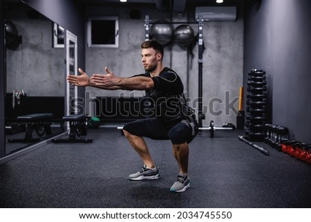 Leg burning exercise, using new EMS technology. A young attractive man in EMS clothes in the gym doing squats with his arms outstretched. Electrical muscle stimulation, strong movement