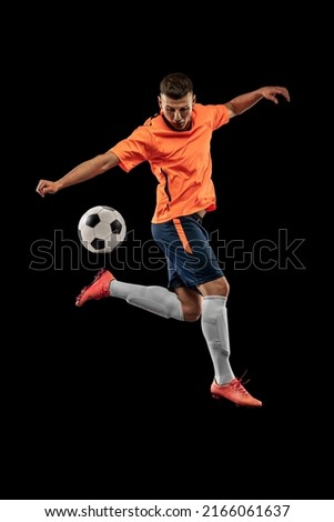 Leg kick. Portrait of professional male football soccer player in motion isolated on dark background. Concept of sport, goals, competition, hobby, ad. Sportsmen wearing orange-blue football kit