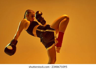 Leg kick. Sportive teen girl, mma fighter athlete in motion, training, fighting against orange studio background in neon lights. Concept of mixed martial arts, sport, hobby, competition, strength, ad