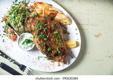 Leg Of Lamb Baked With Sauce In The White Plate Closeup With A Side Dish Of Rice. Rustic Wooden Table And A Glass Of Red Wine.