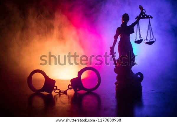 Legal law concept. Silhouette\
of handcuffs with The Statue of Justice on backside with the\
flashing red and blue police lights at foggy background. Selective\
focus