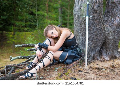 Leggy Medieval Robber Girl Wearing Black Leather Rerebrace, Skirt, Plastron And Vambrace With Iron Estramacon Rests Under The Pine Tree In A Fall Forest