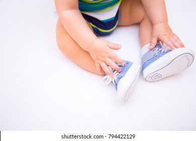 Legs Of Baby Boy Dressed In Children's Blue Sneakers On Lacing On White Background. Concept Of First Shoes.