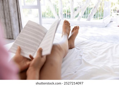 Legs of biracial woman reading book and lying on bed at sunny home. Lifestyle, free time and domestic life, unaltered. - Powered by Shutterstock