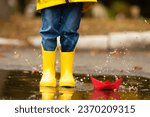 legs of child with rubber boots jump in puddle on an autumn walk. Happy child girl in a yellow jacket and yellow rubber boots has fun with paper boat in a puddle in autumn on nature. Happy childhood