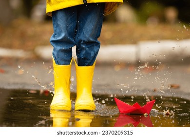 legs of child with rubber boots jump in puddle on an autumn walk. Happy child girl in a yellow jacket and yellow rubber boots has fun with paper boat in a puddle in autumn on nature. Happy childhood