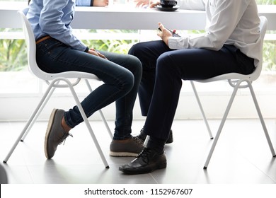 Legs Of Couple Business Sitting On The White Chairs Under The Table Together And Hold Smartphone For Meeting And Drink Coffee In A Black Cup