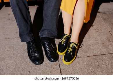 Legs Of A Couple In Love Close Up. Classic Black Men’s Shoes And Playful Women’s Shoes With Yellow Soles And Laces, Close-up Shot Of Shoes.
