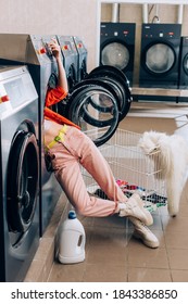 Legs Of Curious Woman Sticking Out Of Washing Machine Near Detergent Bottle And Cart With Dirty Clothing