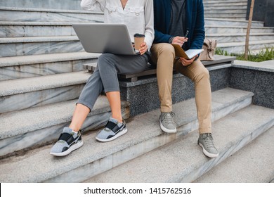 legs in footwear of man and woman sitting on stairs in urban city center in smart casual business style working together on laptop wearing sneakers, stylish freelance people using technology outside