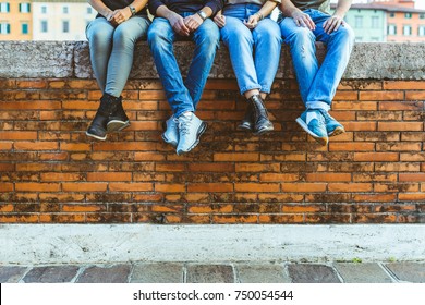 Legs Of Four Teenagers Sitting On A Bricks Wall. Close Up View On Bottom Part Of The Body, Two Girls And Two Boys Sitting Together In The City. Friendship And Lifestyle Concepts