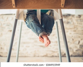 The Legs Of A Little Toddler Sitting In A High Chair At The Dining Table