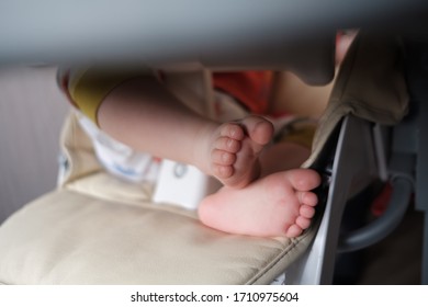 The Legs Of A Little Toddler Sitting In A High Chair At The Dining Table
