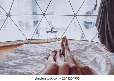 Legs of a man and woman couple in bed against the background of a snow-covered forest in a dome camping. Glamping vacation lifestyle concept.