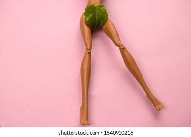 legs of a plastic doll with a green leaf covering the crotch on a pastel pink background