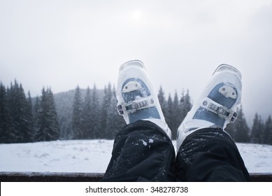 Legs Skier In Ski Boots While Relaxing After A Day Skiing. Mountain Scenery Away