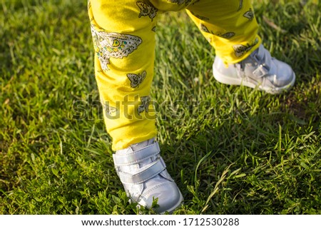 Legs Of Small Baby Girl With Shoes On Young Green Grass. Walk In Summer Park Close-Up.