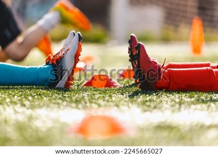 Legs of two soccer players in red and blue soccer socks and cleats sitting on the training ground. Kids on football soccer training pitch. School boys practicing sports on physical education class