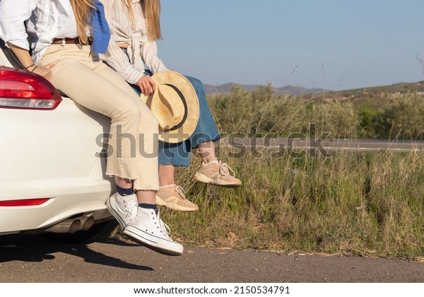 Legs of two unrecognizable\
girl friends sitting in the car truck of the white car admiring the\
sunset