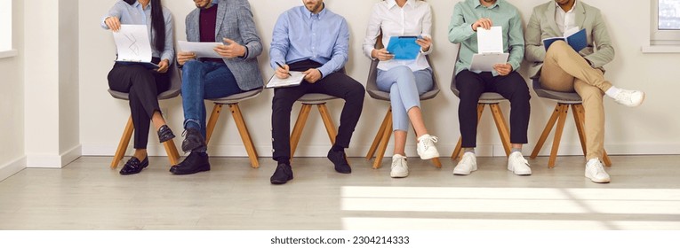 Legs of unrecognizable business people sitting on the chairs in a row with resumes or documents in their hands. Group of a staff. Job candidates seekers waiting for interview invitation turn. Banner. - Shutterstock ID 2304214333
