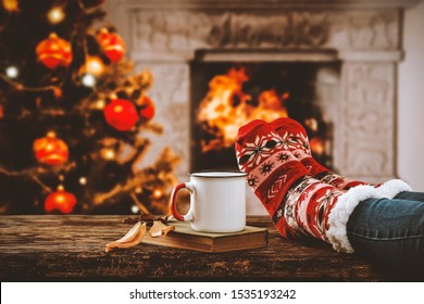 Legs In Winter Christmas Socks On Wooden Top Board With Fireplace Background In Cozy Home Interior.