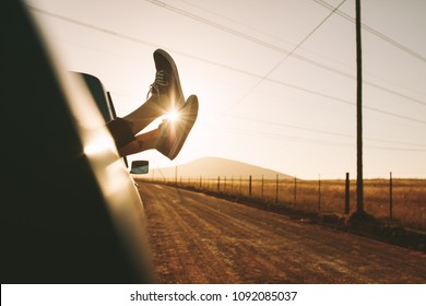 Legs Of A Woman Relaxing At The Back Of A Pick Up Truck On A Highway In Country Side. Legs Hanging Out From Car With Sun In The Background.