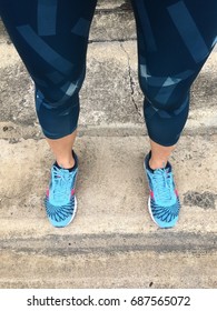 Legs Of A Woman Wearing Blue Training Shoes And Dark Green Patterned Jogging Pants Standing On A Cement Floor