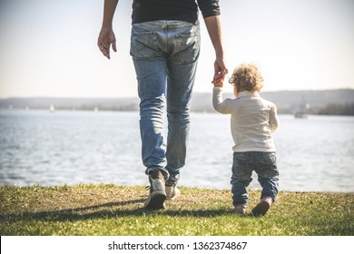 Legs. Young Caucasian father in jeans goes and holds the hand of two year old daughter with his back along the grass near the lake. Dad teaches the child to walk. Father baby spending time together
