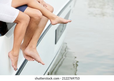 Legs Of Young Couple Sitting On Luxurious Boat And Swinging Legs Over Water