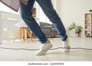 Legs Of Young Man Who, Walking Clumsily, Stumbled Over Electric Cord In Office Or At Home. Man In Sneakers And Jeans Trips Over Black Power Cord Lying On Floor. Low Section.