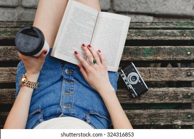 Legs of a young woman in jeans shorts, on a bench in the park. Girl reading a book in a park outdoors with cup of coffee and camera. Close-up. Point of view