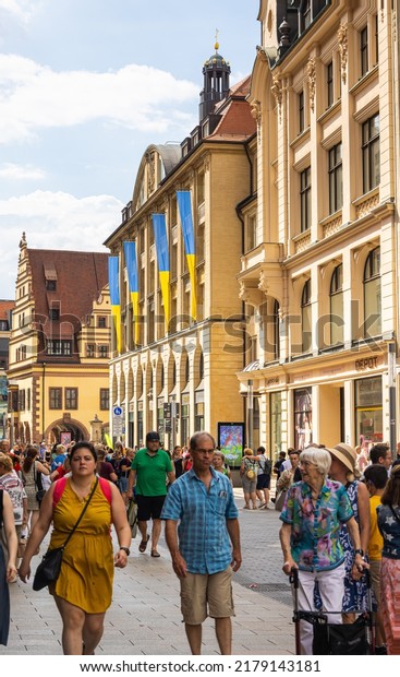 Leipzig, Germany - June 25, 2022: Street view\
into the Grimmaische Strasse. Tourists and locals stroll across the\
street with many shops. Blue and yellow flags on the houses (city\
Color of Leipzig)