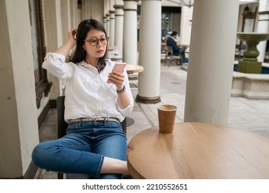 Leisure Asian Business Lady Wearing Earphones Binge Watching Online Tv Shows On Phone During Lunch Break At Cafe In Palo Alto City California Usa. She Folds Legs And Scratches Head While Watching