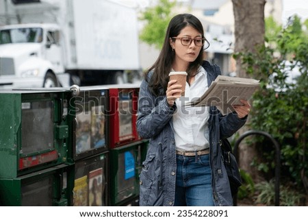 leisure asian Korean businesswoman in smart casual reading tabloid by newspaper boxes on the street and drinking daily coffee in the morning at downtown palo alto California usa