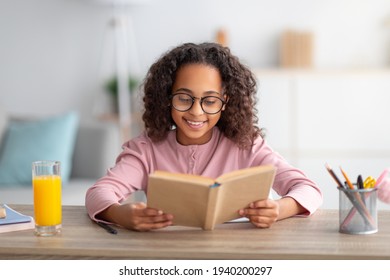 Leisure Concept. Cute Black Schoolgirl Reading Paper Book And Drinking Orange Juice, Sitting At Desk