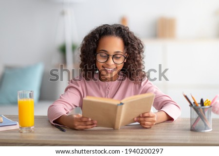 Leisure concept. Cute black schoolgirl reading paper book and drinking orange juice, sitting at desk