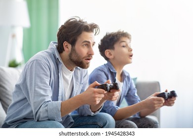 Leisure With Dad. Concentrated Father And Son Competing With Each Other In Online Video Games, Using Joysticks, Having Fun At Home. Family Sitting On Sofa And Enjoying Time Together