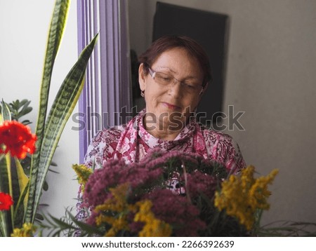 Leisure of the elderly. An older woman is at home, stands near the window and looks with a smile at the autumn bouquet of flowers. Call your elderly family and brighten up their leisure time.
