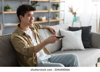 Leisure And Entertainment Concept. Smiling Asian Guy Watching TV Show Or Film, Holding Remote Control. Young Casual Man Enjoying Free Time Sitting On The Comfortable Couch At Home In Living Room