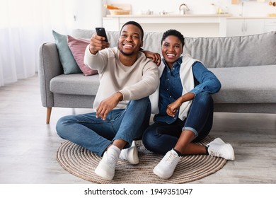 Leisure And Entertainment. Smiling African American couple watching TV show or film, guy holding remote control. Young man and woman enjoying free time sitting on floor carpet at home in living room