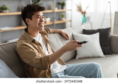 Leisure And Entertainment. Smiling Asian Guy Watching TV Show Or Film, Holding Remote Control. Young Man Enjoying Free Time Sitting On The Comfortable Couch At Home In Living Room, Free Space