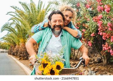 Leisure Fun Activity For Adult Young Caucasian Couple Having Fun Together On A Bike. Man Carry Woman And Ride Bycicle On The Road. Flowers And Spring Nature In Background. People In Friendship