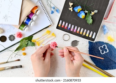 Leisure. Hobbies and recreation. Creating flowers and ornaments from cold porcelain. Painting of jewelry. Woman's hands hold flower made of cold porcelain and brush against background of materials